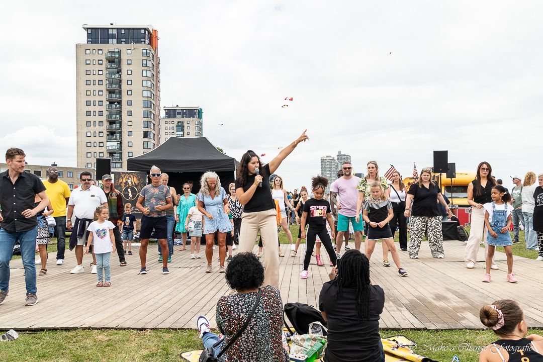 Een groep mensen danst op een houten platform buiten, met een vrouw die de dans leidt en hoge gebouwen op de achtergrond.