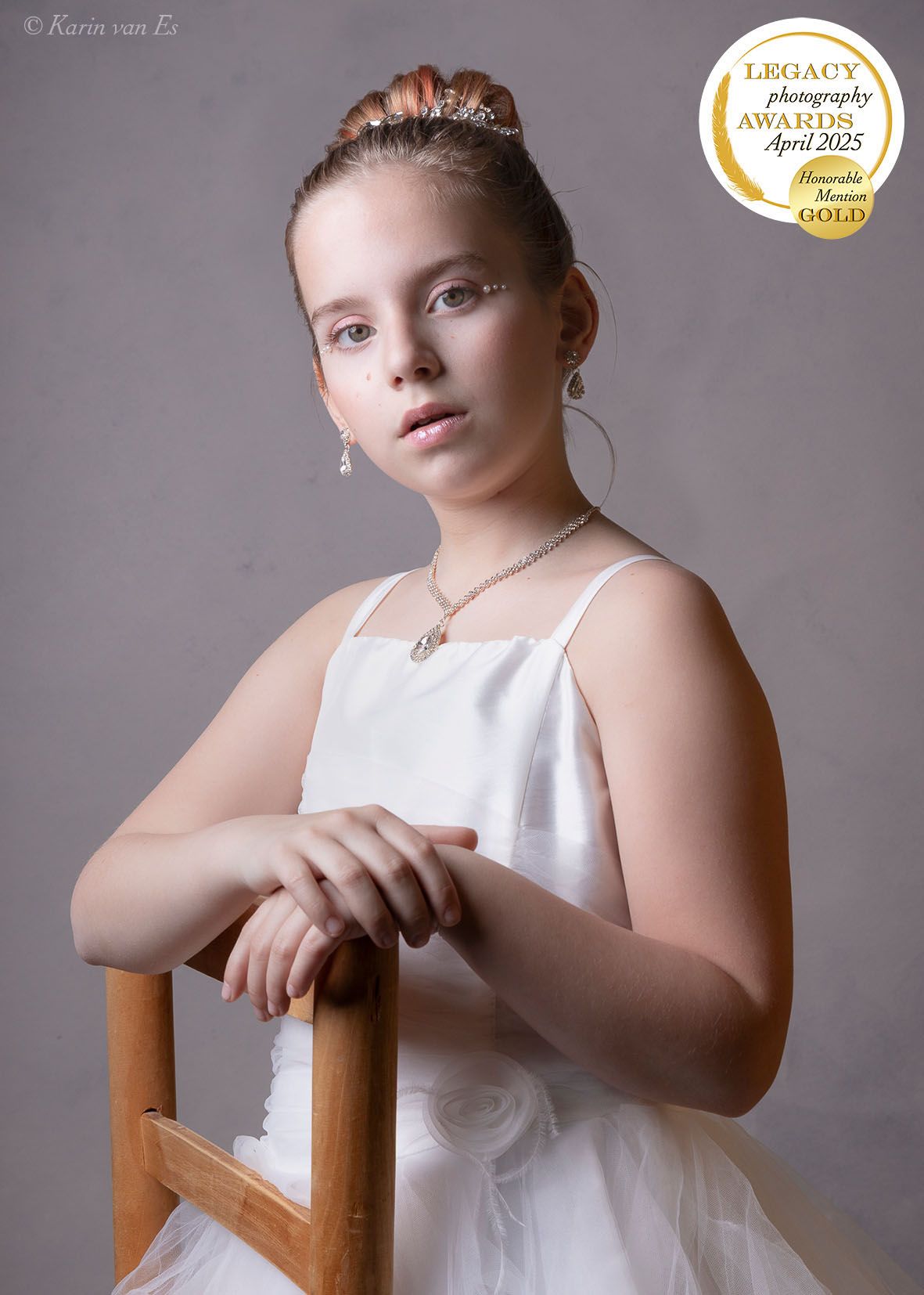 Jong meisje met opgestoken haar en witte jurk poseert, leunend op een houten stoel. Ze draagt sieraden en heeft een serieuze uitdrukking.