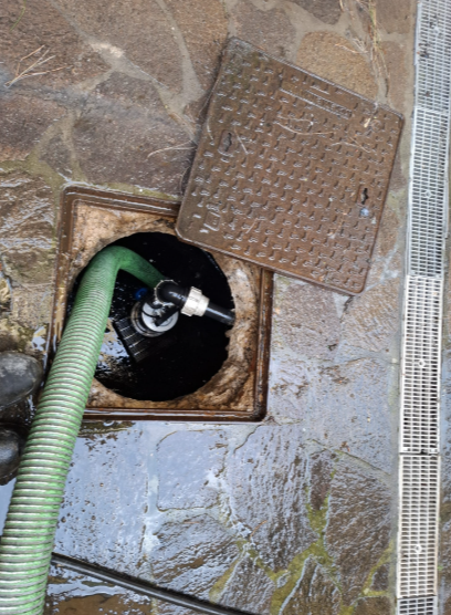 Canale di scolo a vista con tubo verde e coperchio metallico su pavimentazione in pietra bagnata.