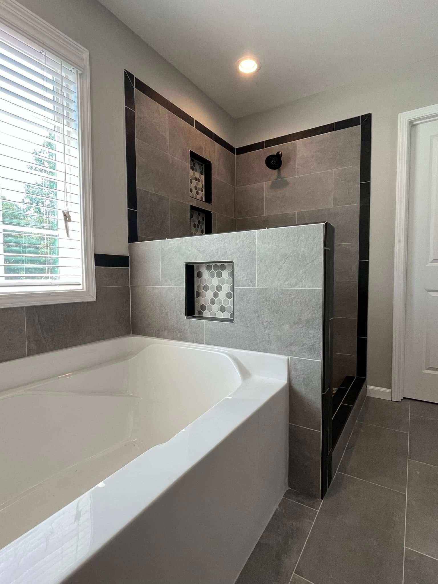 Bathroom with gray tiled shower, built-in tub, white door, and window with blinds.