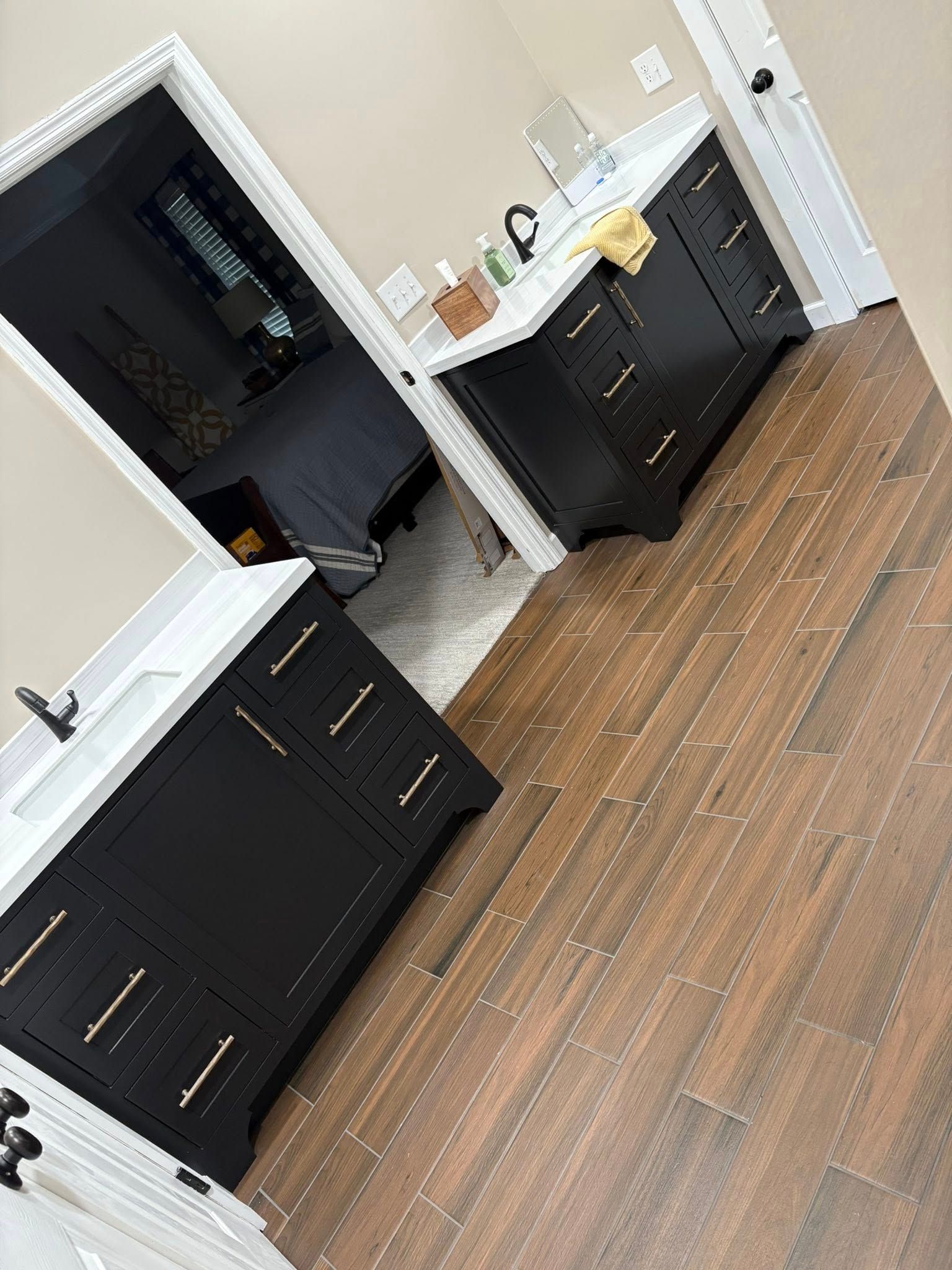 Bathroom with black vanities, white countertops, brown wood-look floors. Doorway to bedroom.