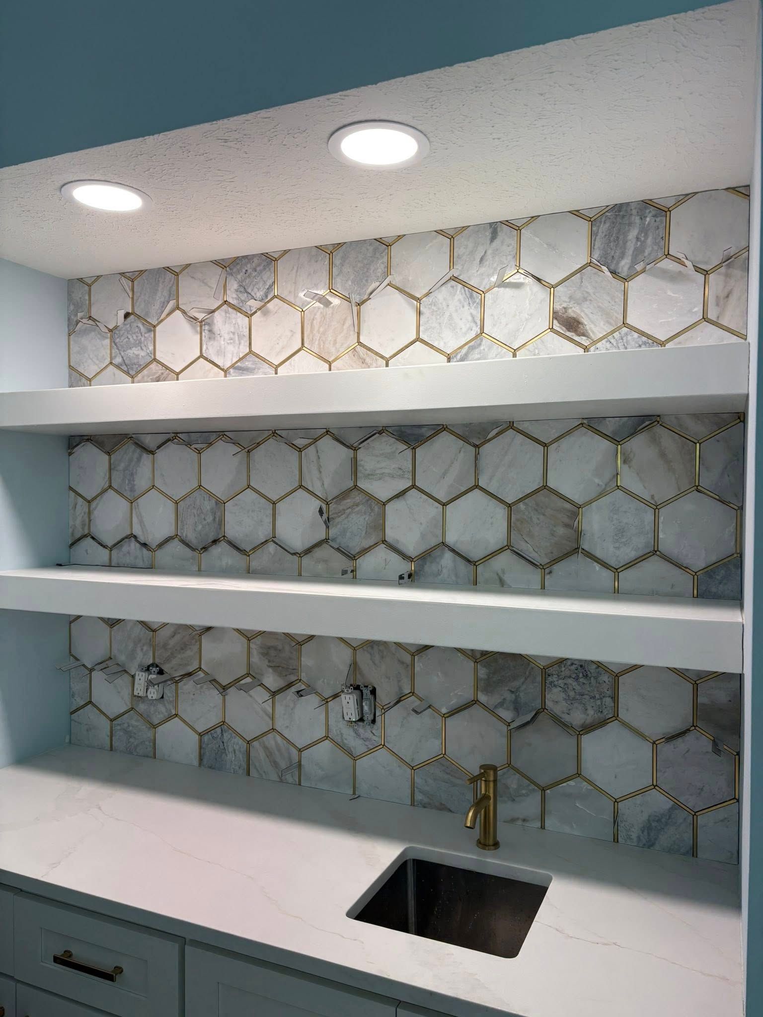 Bar area with hexagon-tiled backsplash, white shelves, and sink, lit by recessed lighting.