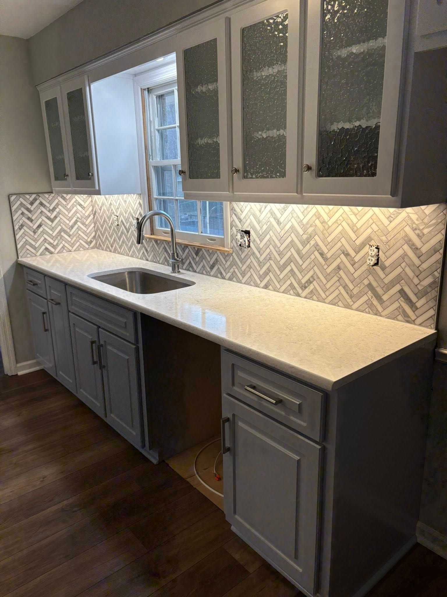 Gray kitchen with countertop, cabinets, sink, patterned backsplash, and window.