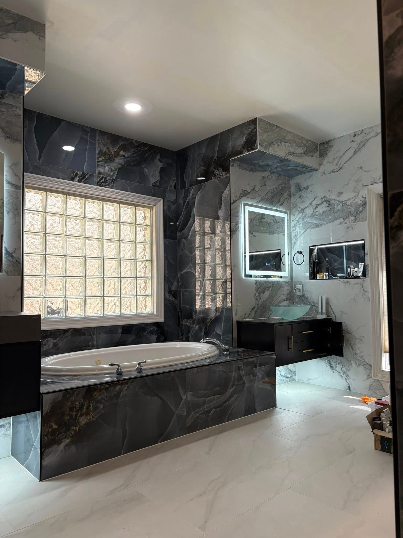 Luxurious bathroom with marble-like walls and floor, dark-colored tub, and lit vanity mirror.