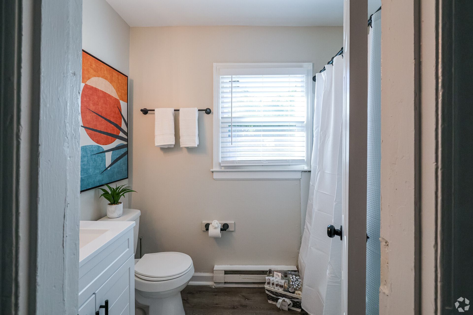 Bathroom with toilet, artwork, towels, shower curtain, and window with blinds.