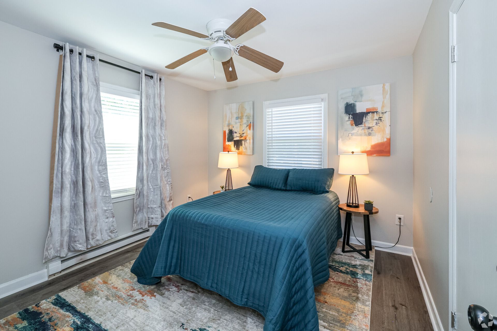 Bedroom with blue bedspread, art, window, rug, ceiling fan, and bedside tables.