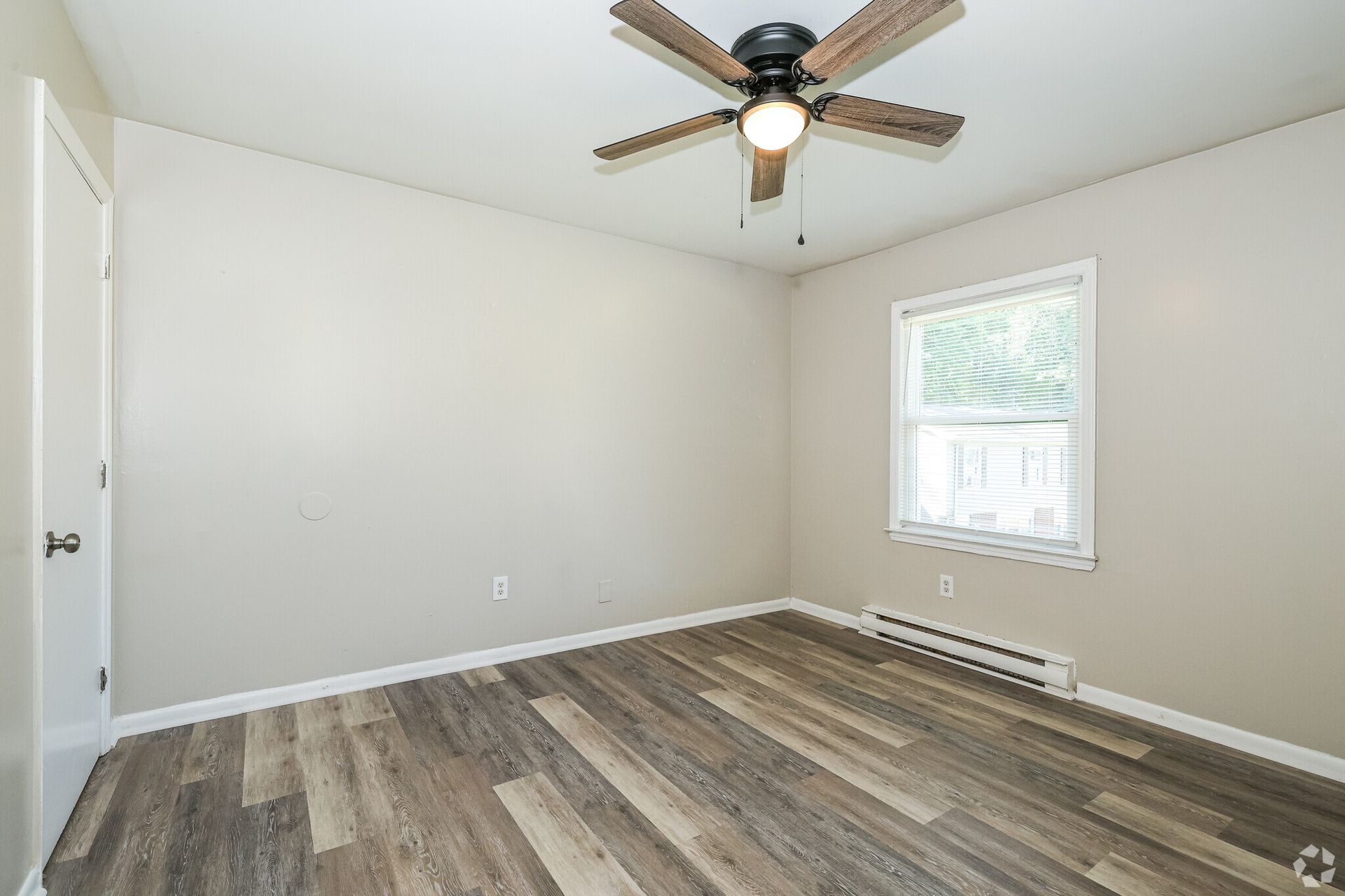 Empty bedroom with wood-look flooring, tan walls, window with blinds, ceiling fan, and door.