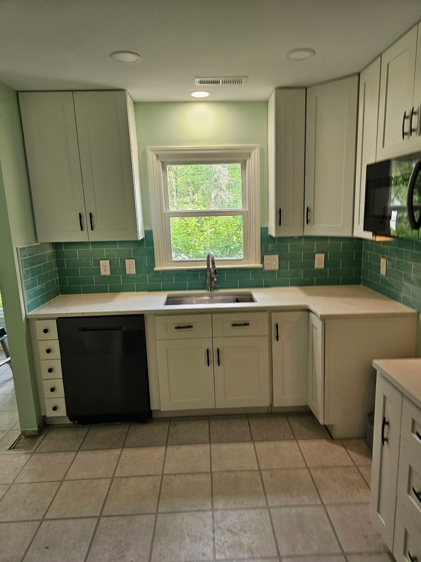 Kitchen with white cabinets, light countertops, a dark dishwasher, and a green subway tile backsplash by a window.