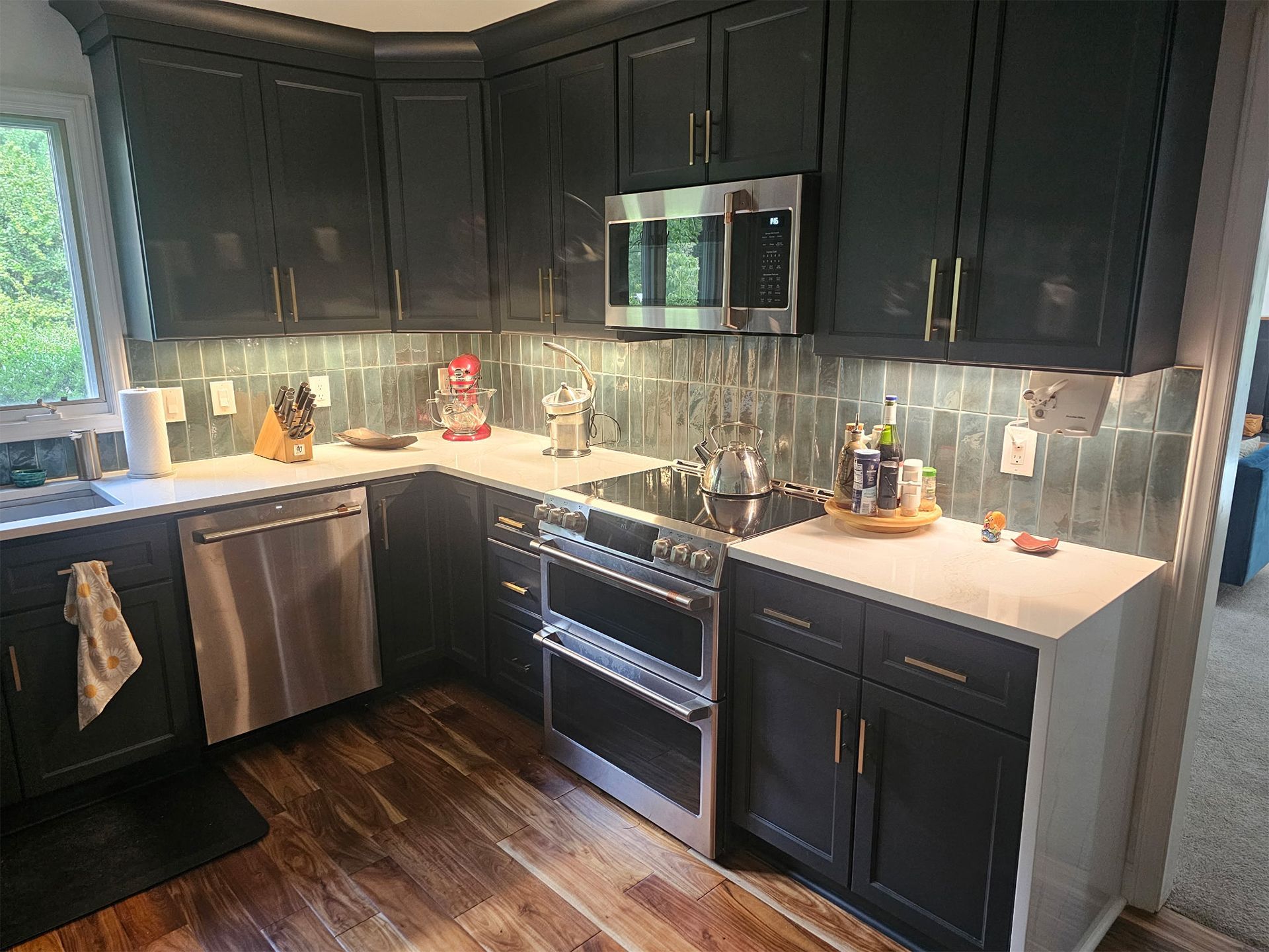 A modern kitchen with dark cabinets, stainless steel appliances, white countertops, and a textured green tile backsplash.