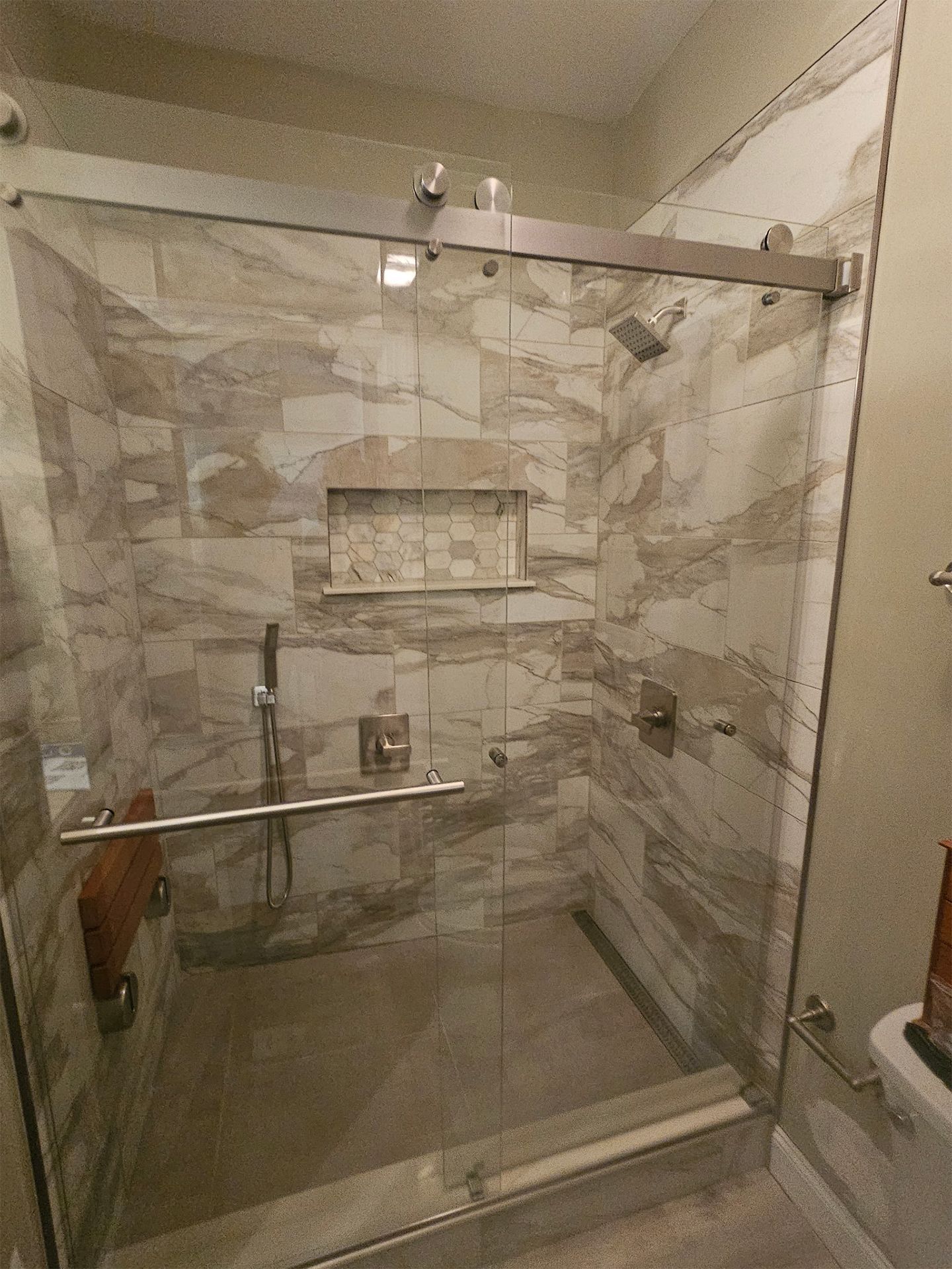 A walk-in shower with a glass sliding door, beige marbled tile walls, a built-in shelf, grab bars, and a wooden bench.