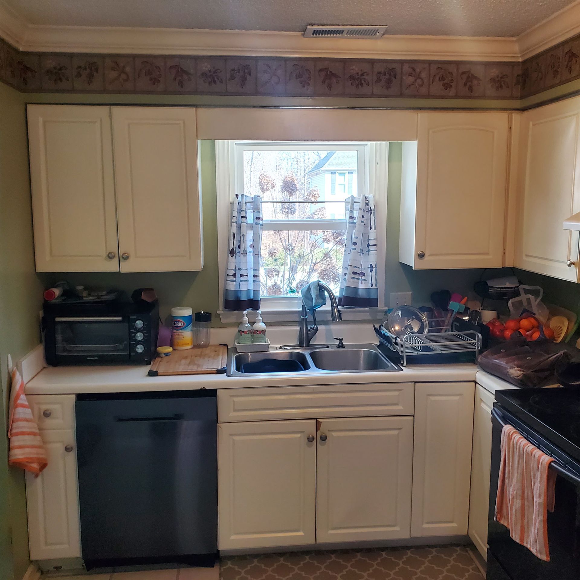 A kitchen sink area with off-white cabinets, a stainless steel dishwasher, a window with curtains, and dark countertops.