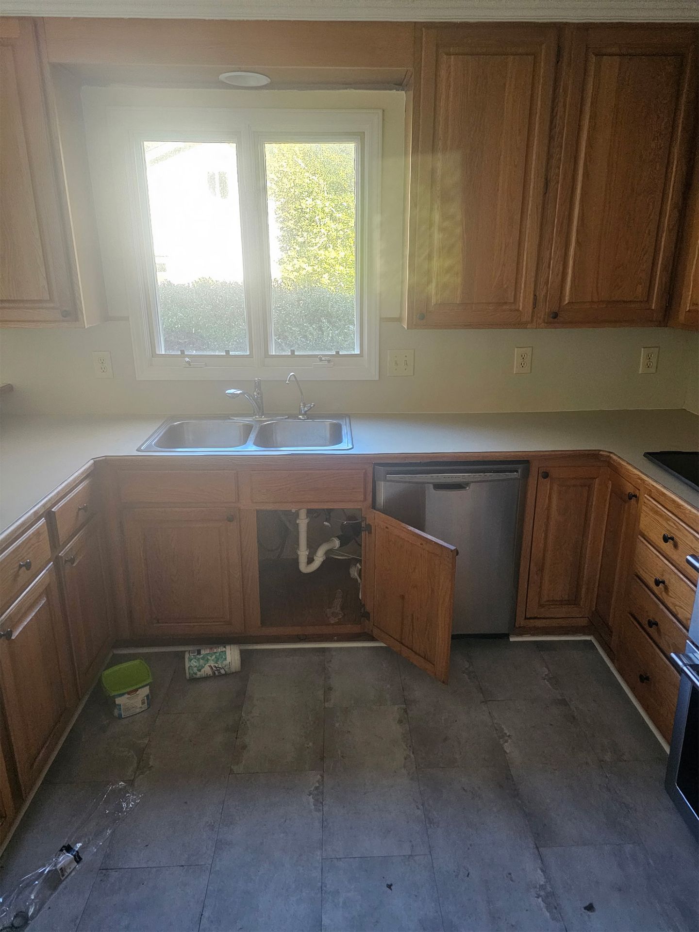 A kitchen with wooden cabinets, a sink, a dishwasher with an open door, and gray flooring beneath a window.