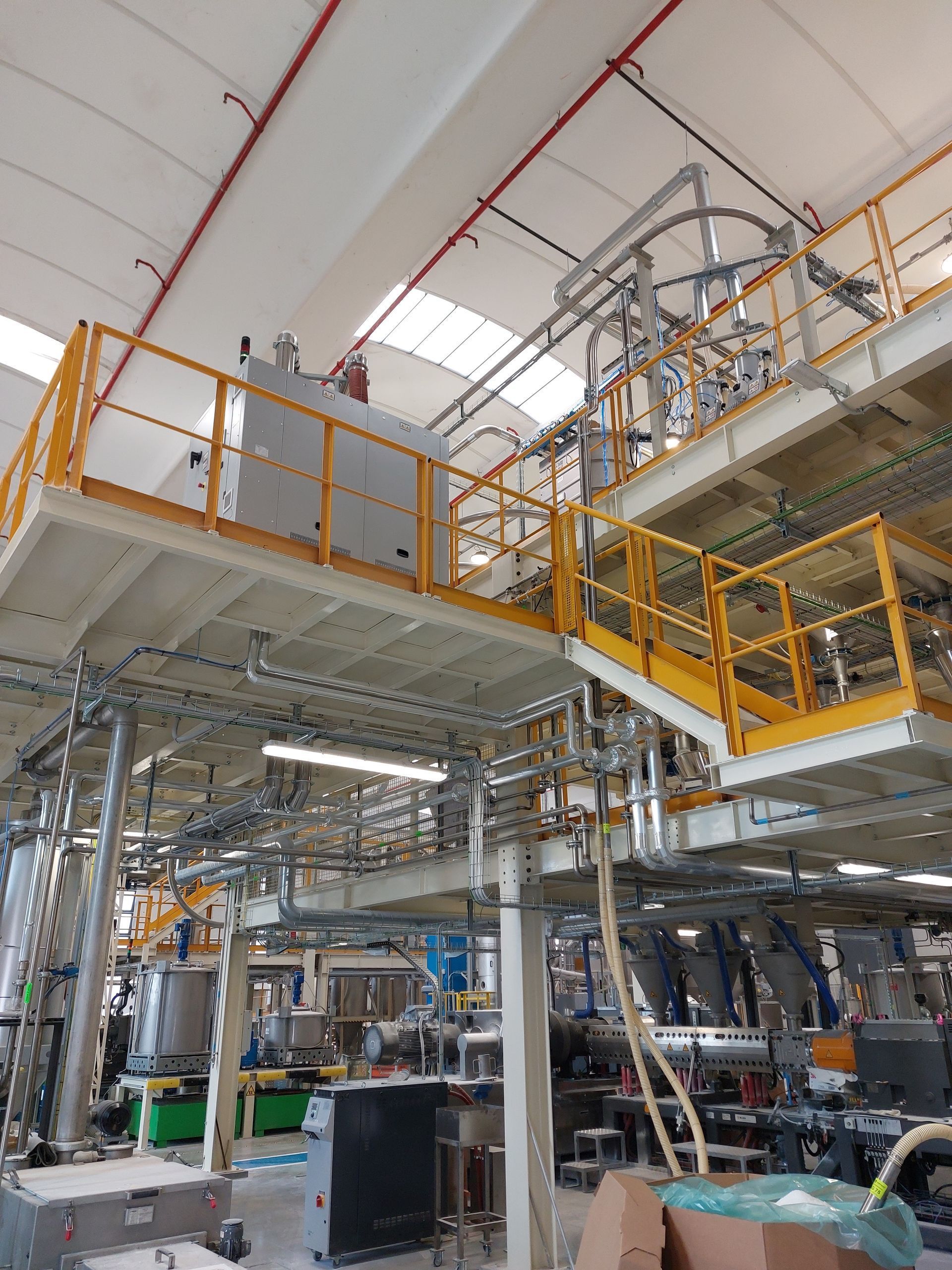 Interno di una fabbrica industriale con passerelle gialle e macchinari in acciaio sotto un alto soffitto bianco.