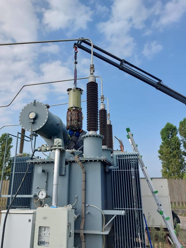 Operai che effettuano la manutenzione di un grande trasformatore elettrico con una gru in una sottostazione all'aperto.