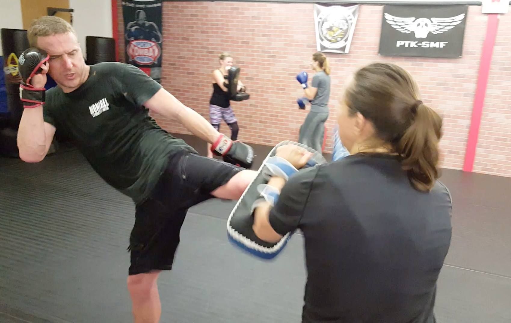 a man is kicking a shield in a boxing ring while a woman watches .