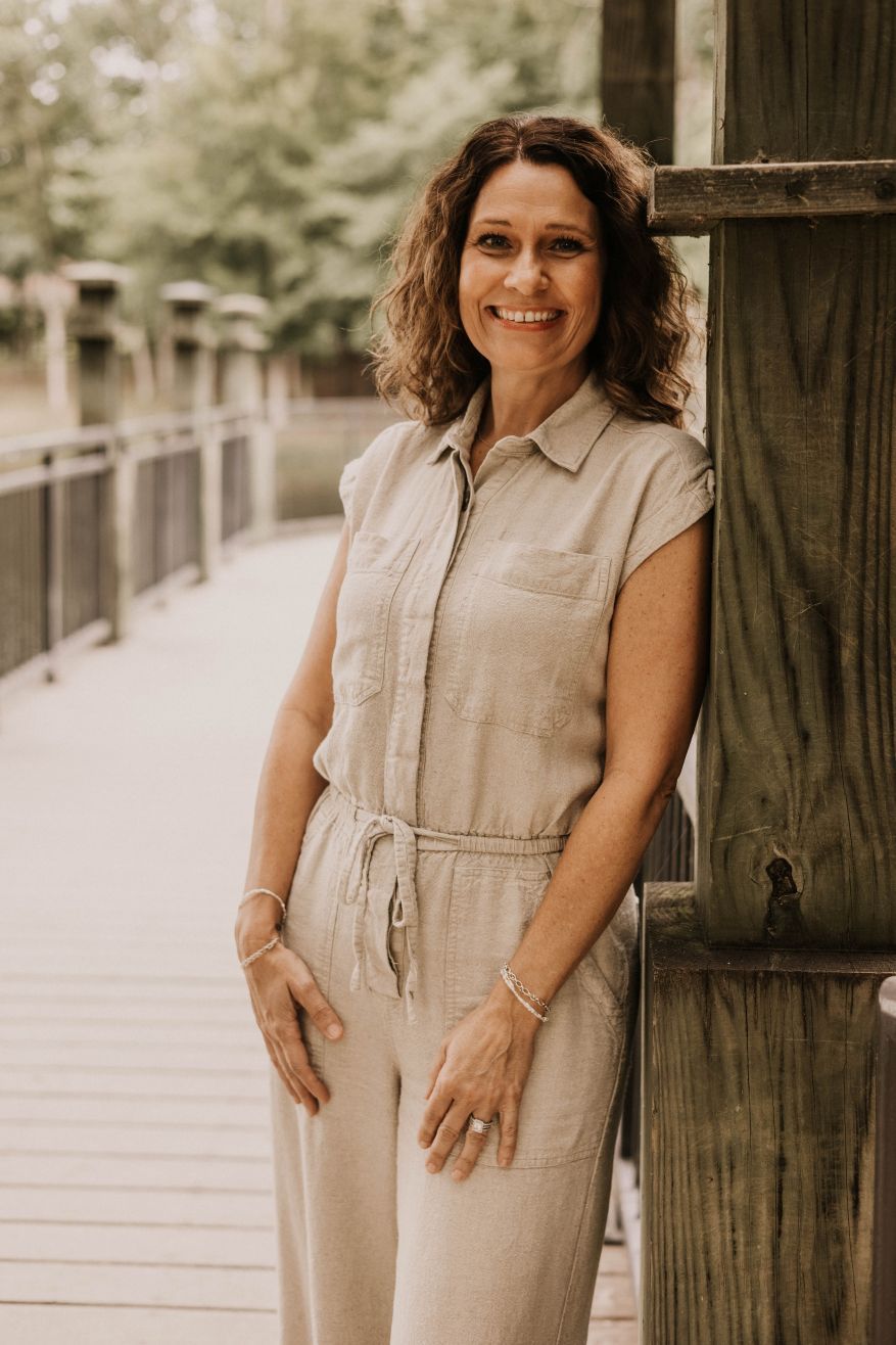 Woman in beige jumpsuit smiles, leaning on wooden bridge rail outdoors.