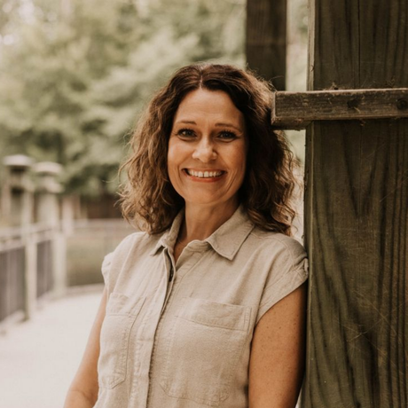 Woman in tan jumpsuit smiles, leaning on wooden structure of a bridge. Outdoors, trees visible.