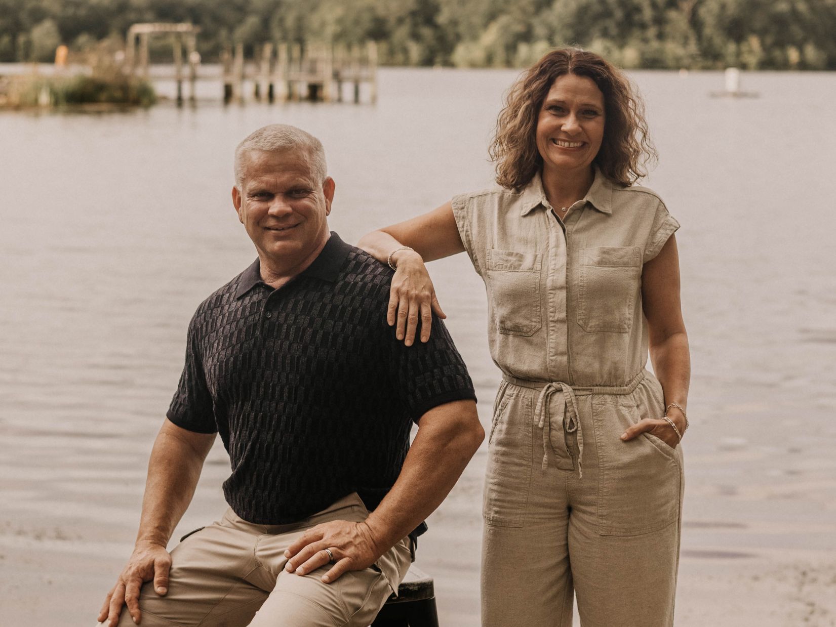 Man and woman by lake; man seated, wearing black shirt and khaki pants; woman standing in tan jumpsuit.