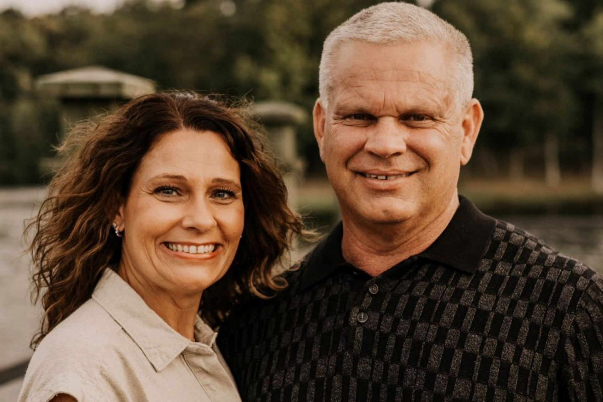 Smiling couple outdoors, man in black polo shirt, woman in tan jumpsuit, lake and trees in background.
