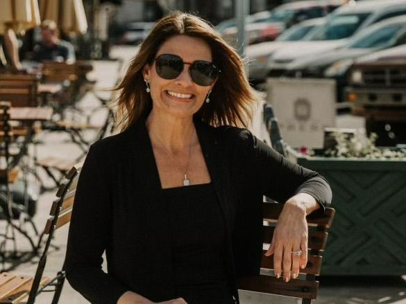 A woman wearing sunglasses and a black dress is leaning on a wooden bench.