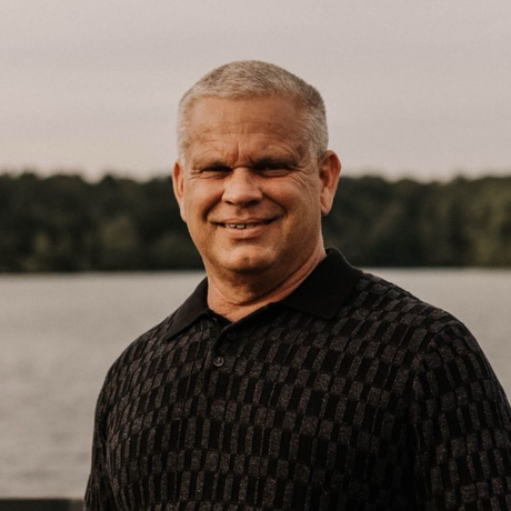 Man with gray hair smiling outdoors, wearing a black patterned shirt, lake in background.