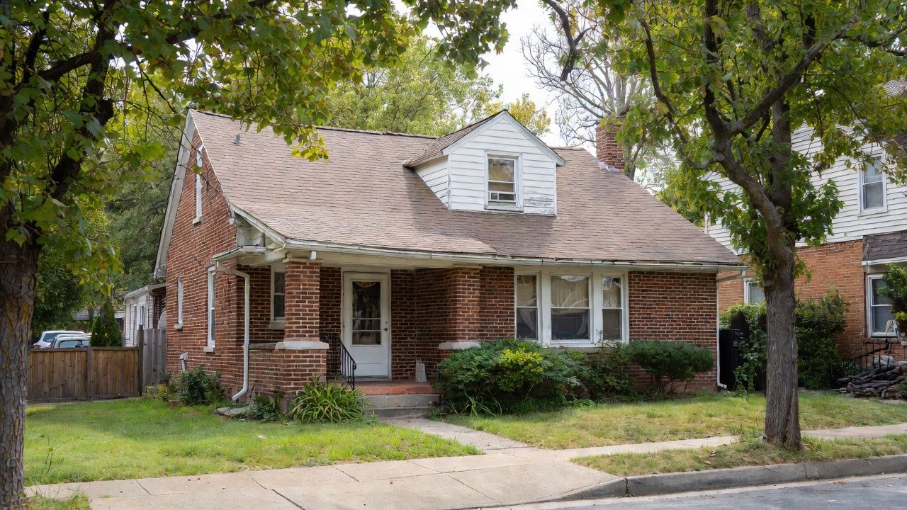 Brick suburban house with a covered porch, white dormer, and large trees in front