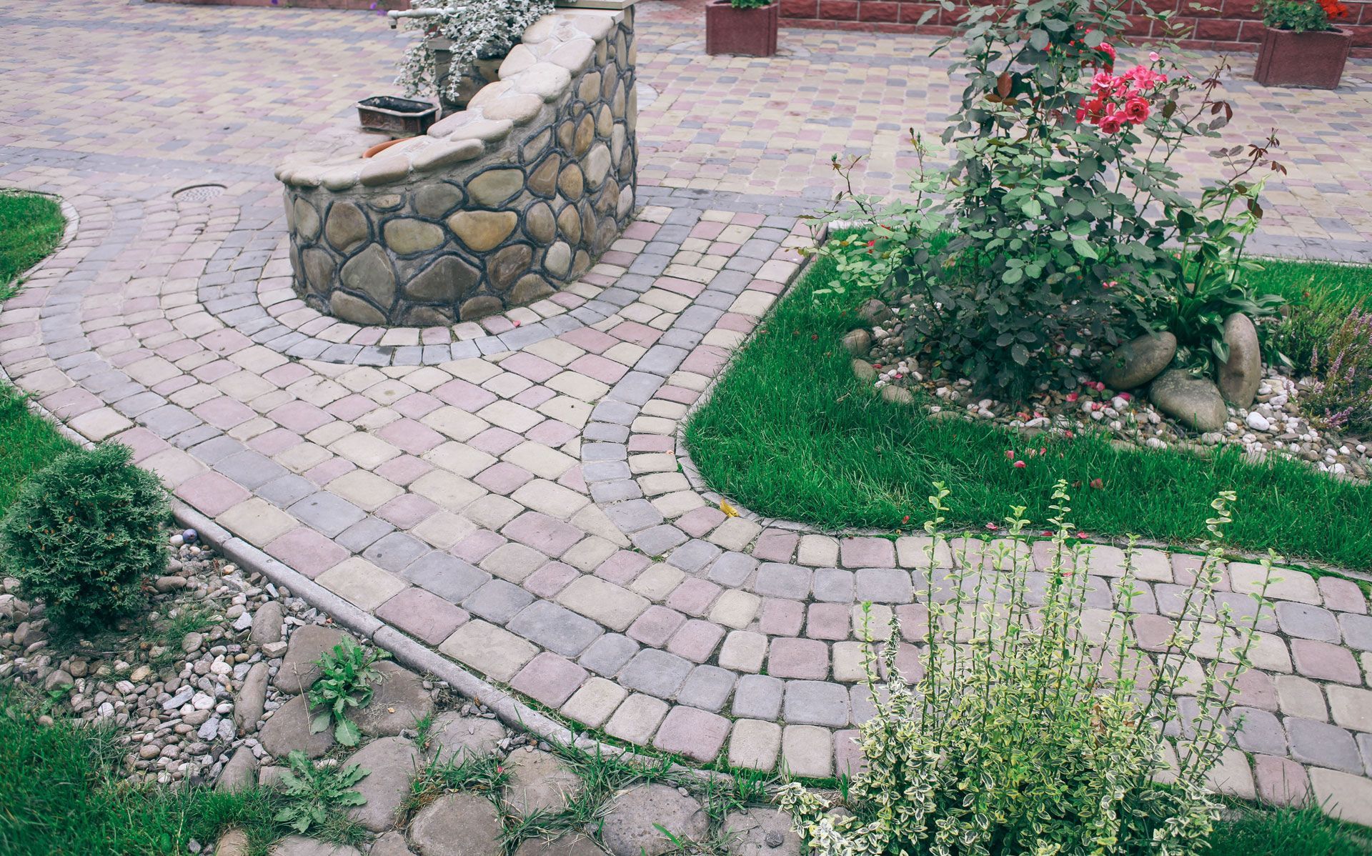Curving brick pathway through a garden with a stone wall and flowerbeds, green grass.