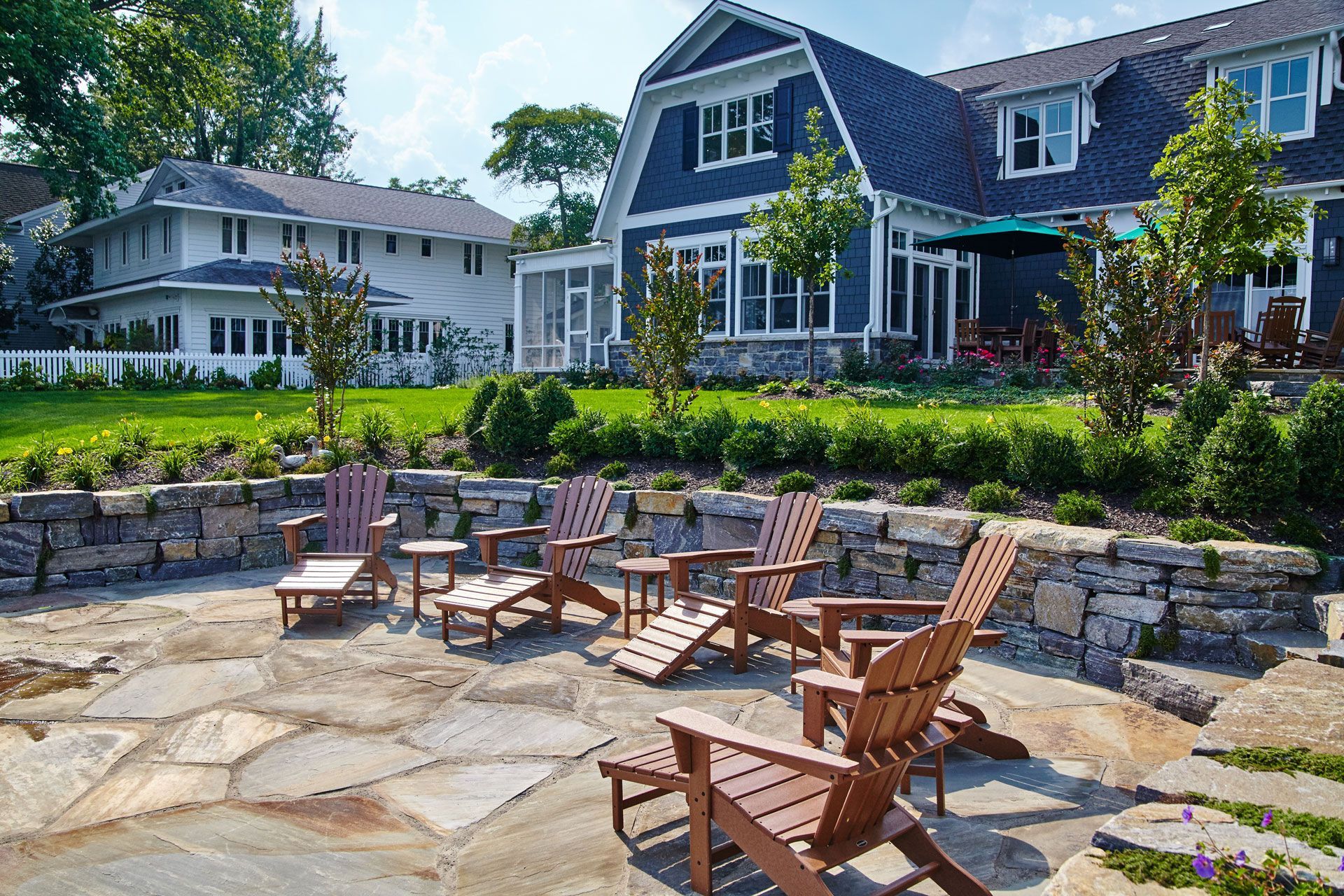 Patio with wooden chairs and stone wall, overlooking a blue house and lawn.