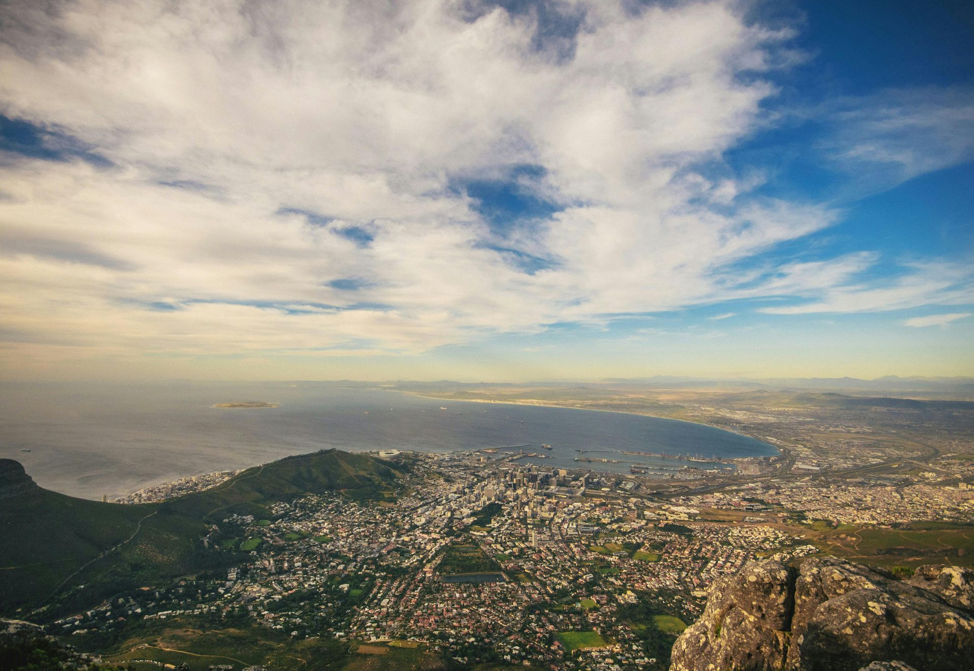 View of Cape Town, South Africa, from a mountaintop, featuring city, sea, and partly cloudy blue sky.