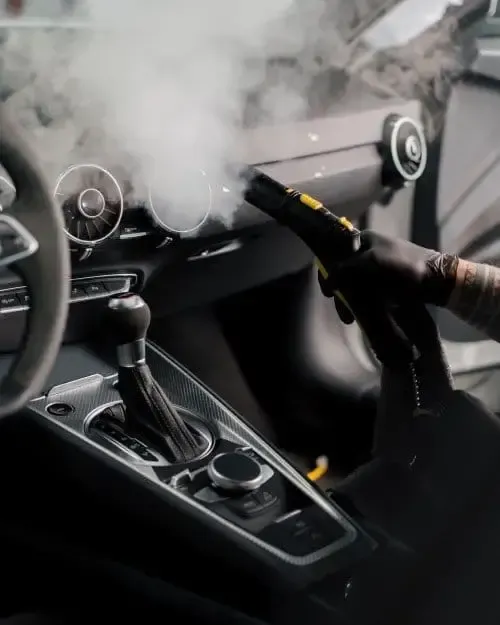 Car interior being steam cleaned; hand in glove holding a steam cleaner, visible steam rising.