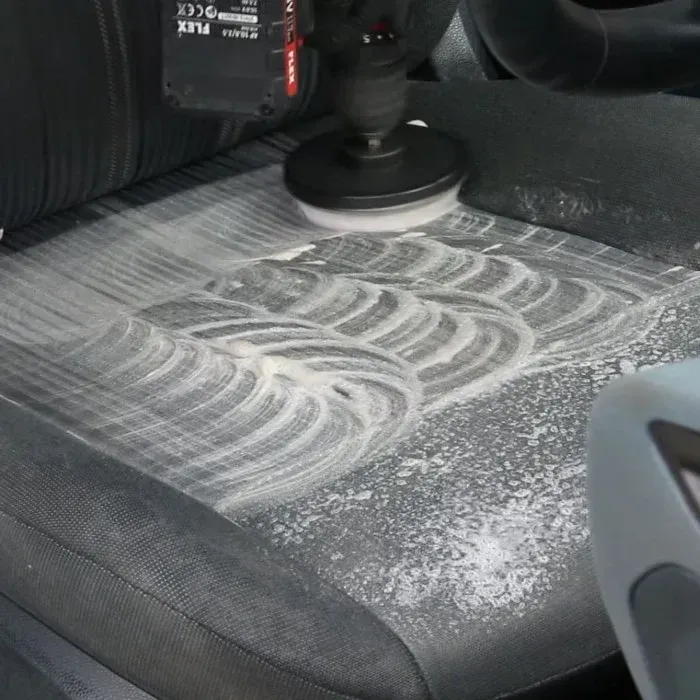 A power drill with a brush cleans a car seat covered in fabric shampoo for automobiles.