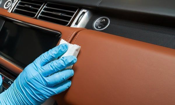 A gloved hand wipes a tan leather car dashboard with a white cloth, applying interior ceramic coating.