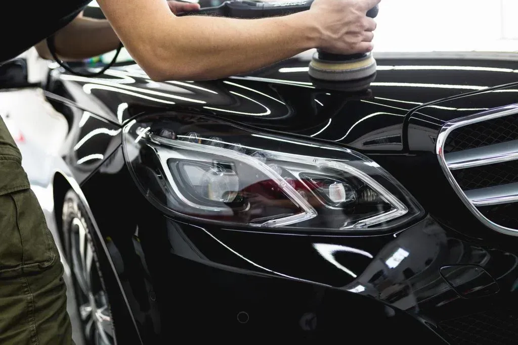 Luxline employee does a 2 step polish on the black hood of a Mercedes Benz with a buffer in a well-lit garage.