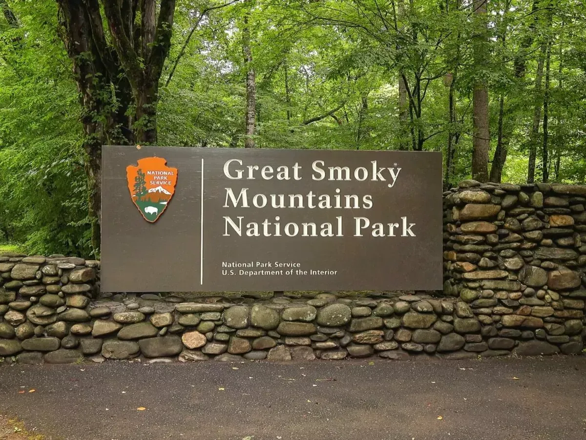 A brown Great Smoky Mountains National Park sign with a stone wall base, set against a background of green trees.