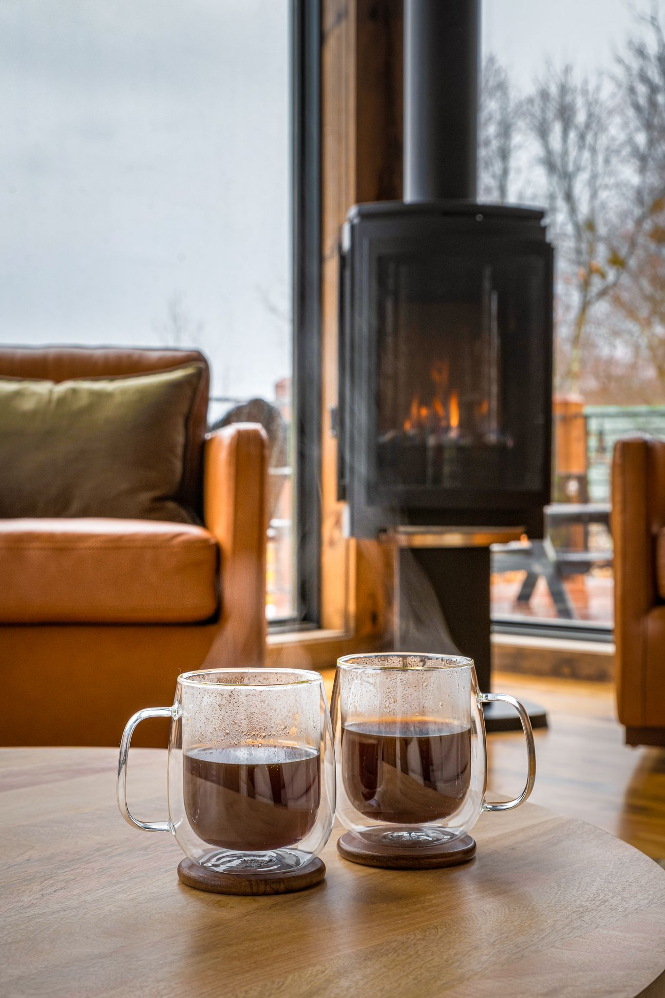 Two glass mugs of dark coffee sit on a wooden table in a cozy living room with a lit fireplace and leather armchairs.