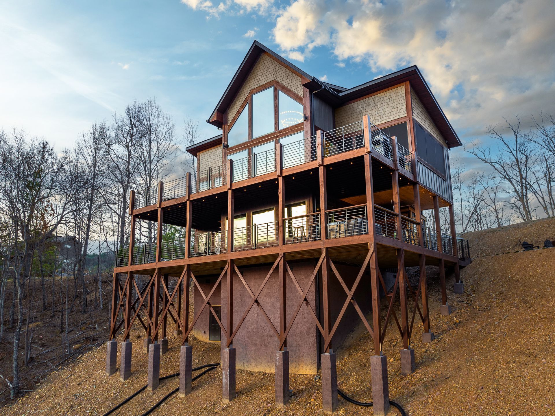 A multi-level cabin house on stilts sits on a dry, grassy hillside under a partly cloudy sky.