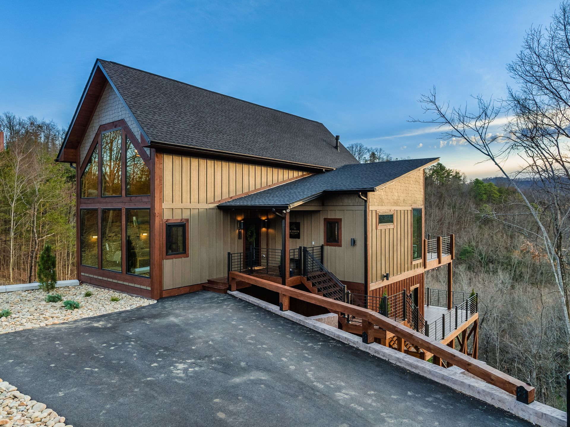 A tan, modern rustic cabin sits on a hillside with a dark asphalt driveway, large windows, and a tiered wooden deck.