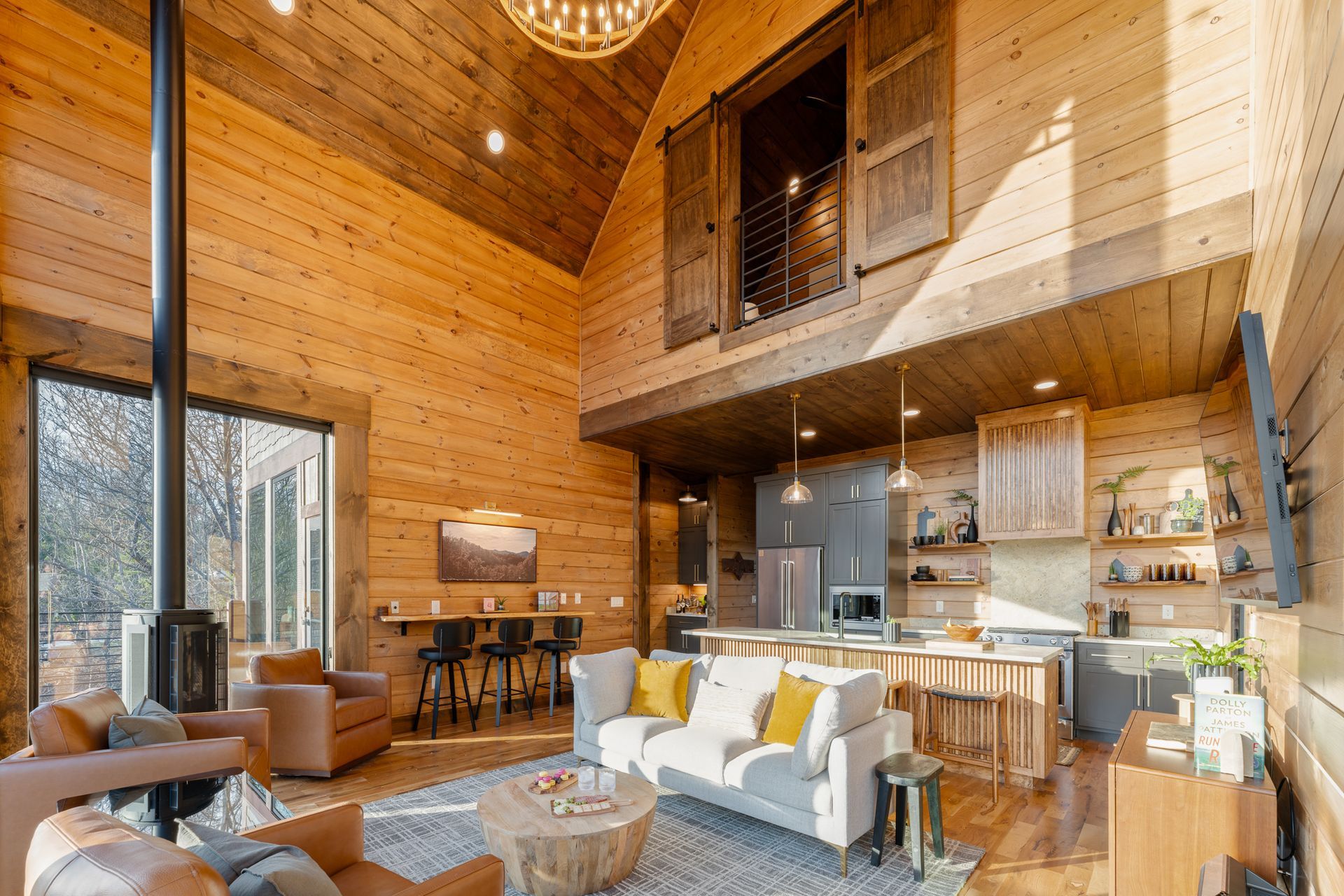 Rustic cabin interior with wood-paneled walls, a high ceiling, a kitchen, living area, and loft space.