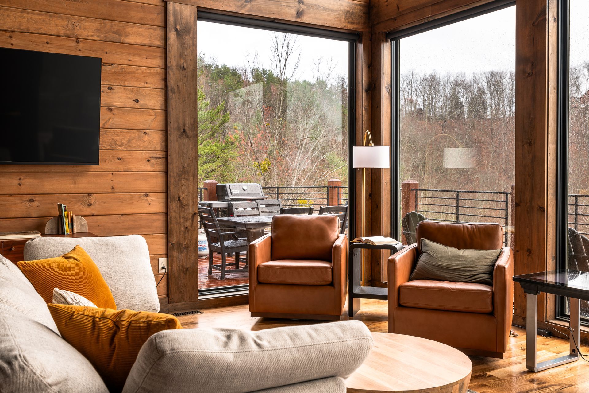 A cozy living room with a light-colored sofa, two leather armchairs, and floor-to-ceiling windows overlooking a deck.