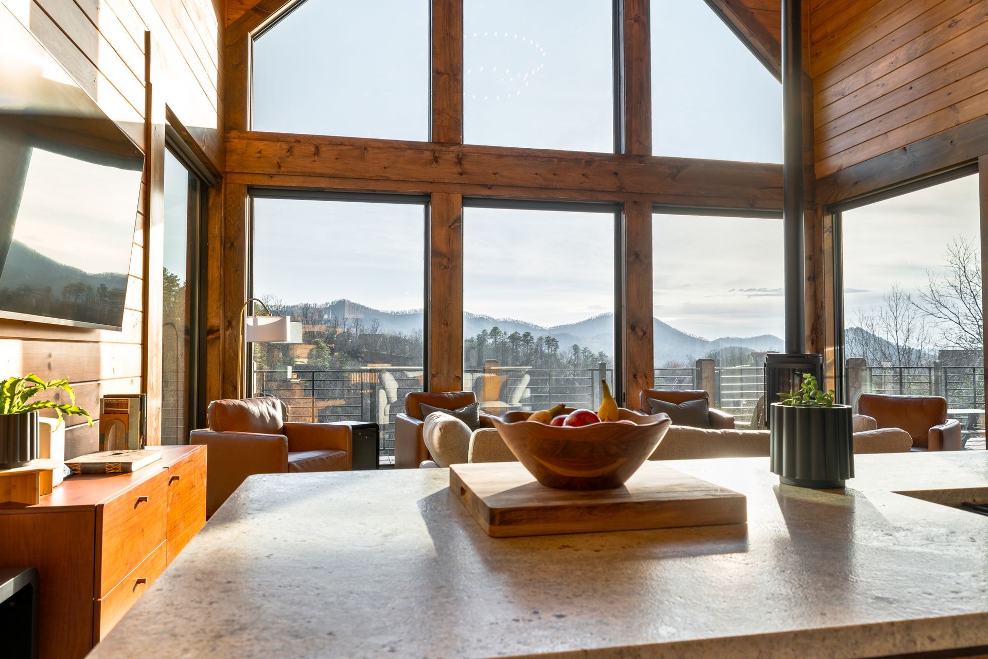 A modern, wood-framed cabin interior features a stone kitchen island, a wooden bowl of fruit, and floor-to-ceiling windows.
