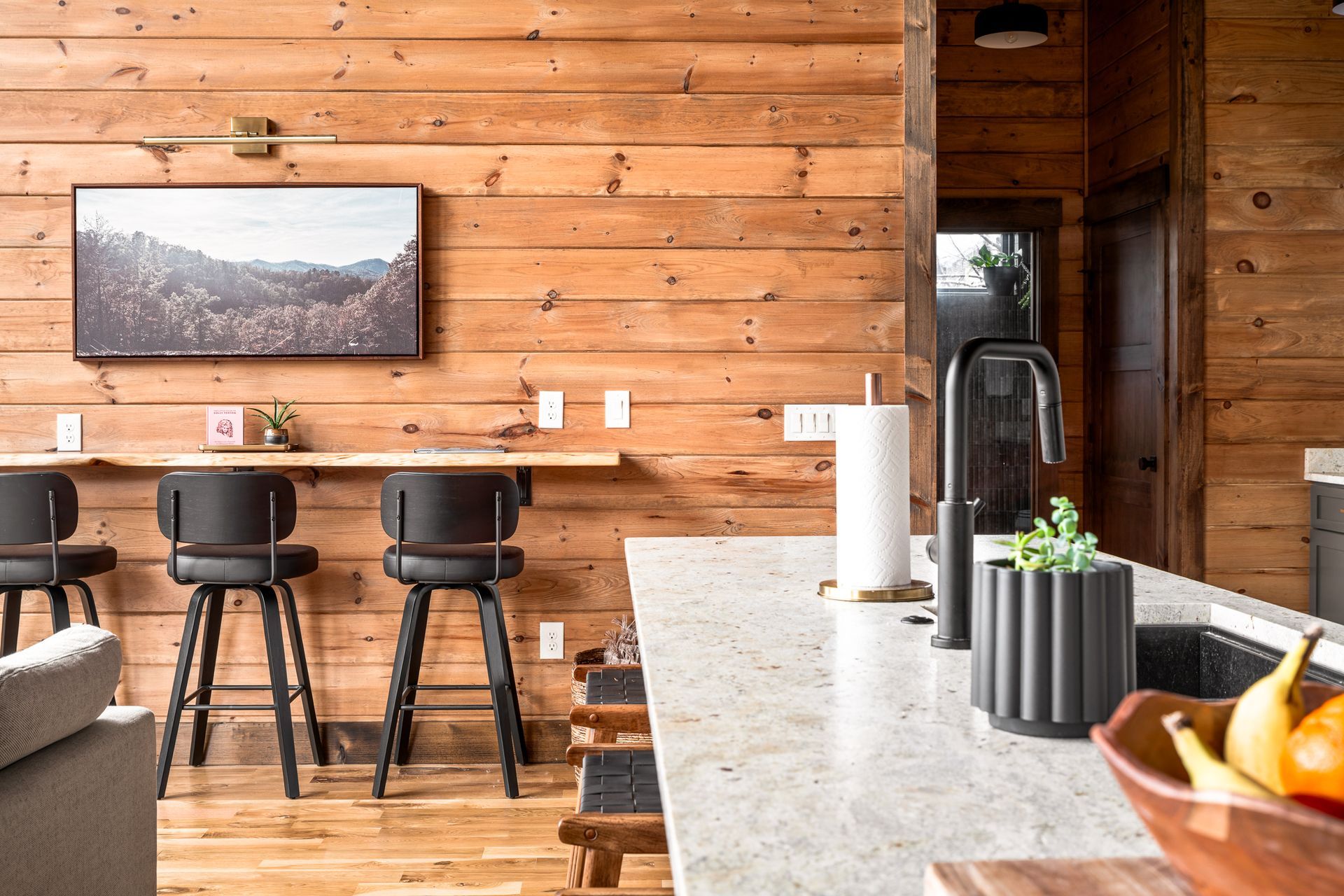 Modern rustic kitchen with wood-paneled walls, a marble island countertop, black stools, and a small plant in a planter.