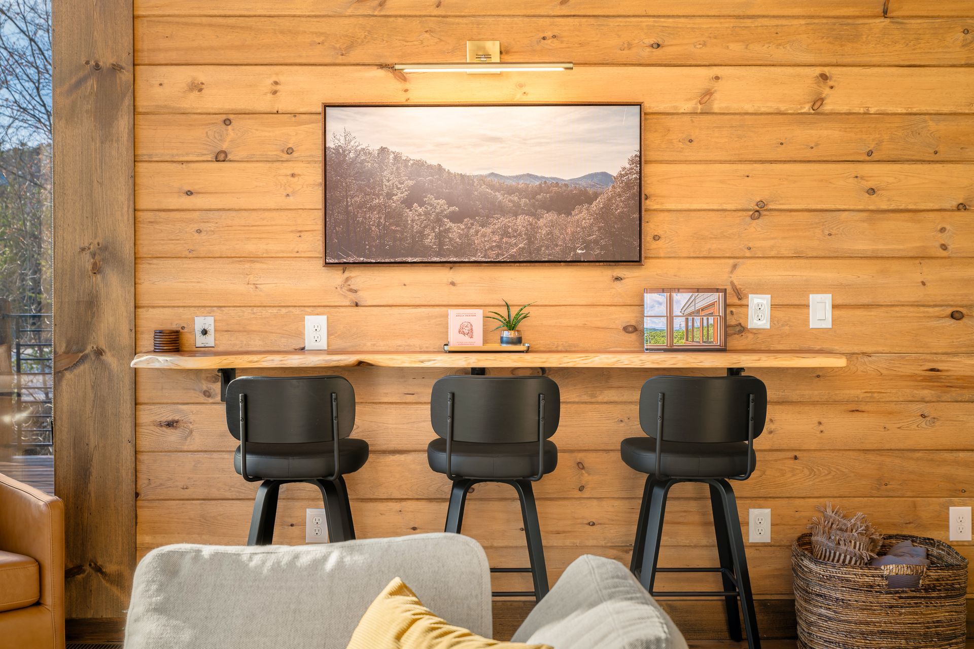 Three black bar stools placed at a wooden wall-mounted counter, with landscape art, plants, and decor on the wall.