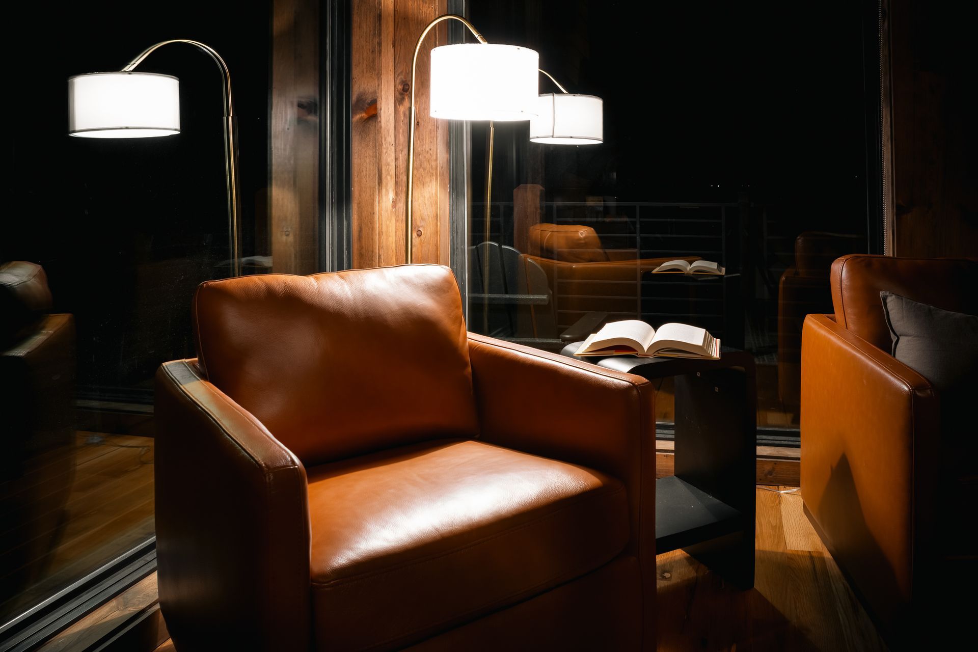 A brown leather armchair with an open book on a side table, illuminated by two modern floor lamps in a dimly lit room.