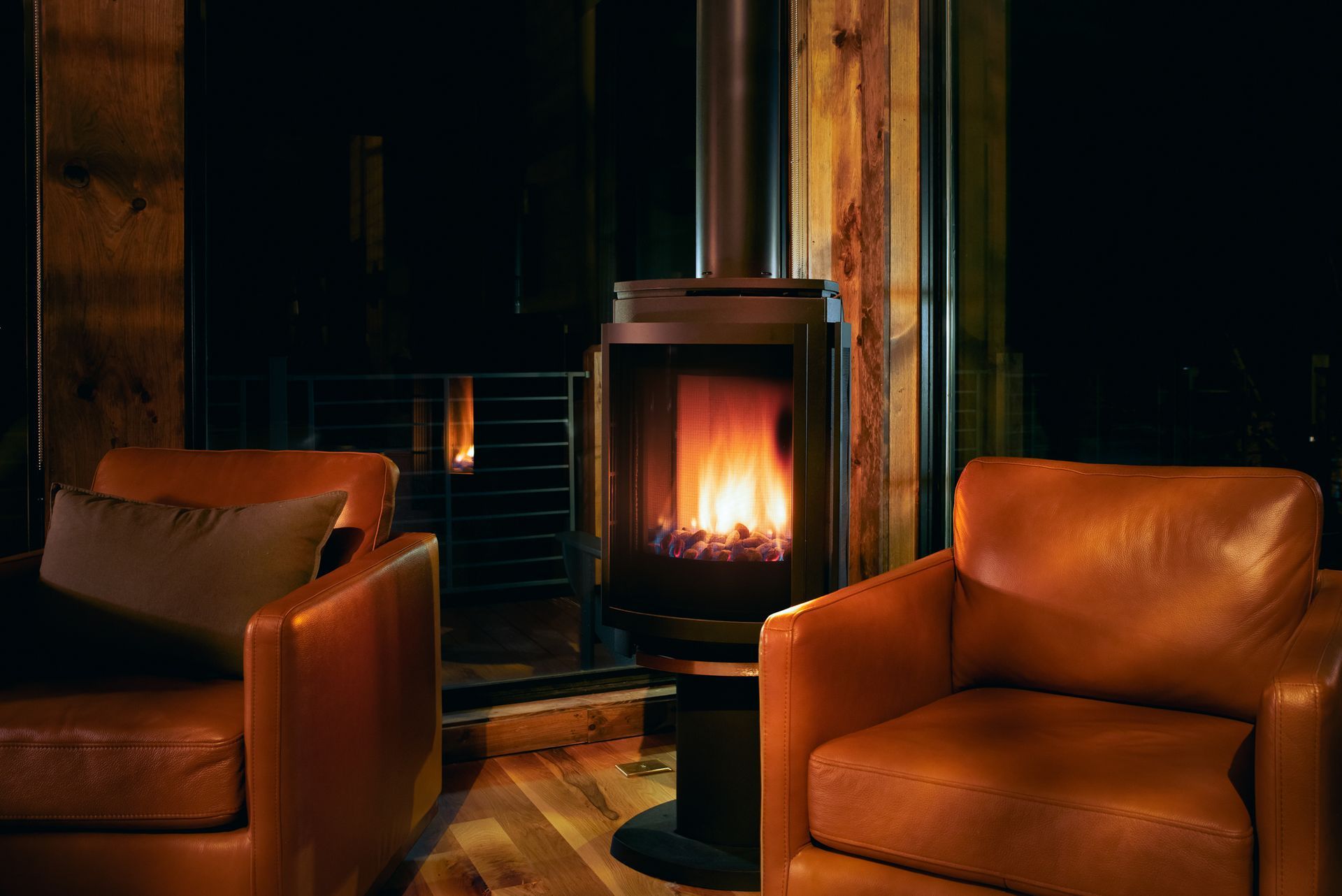 A lit fireplace sits between two tan leather armchairs in a dimly lit, rustic room with wooden walls.