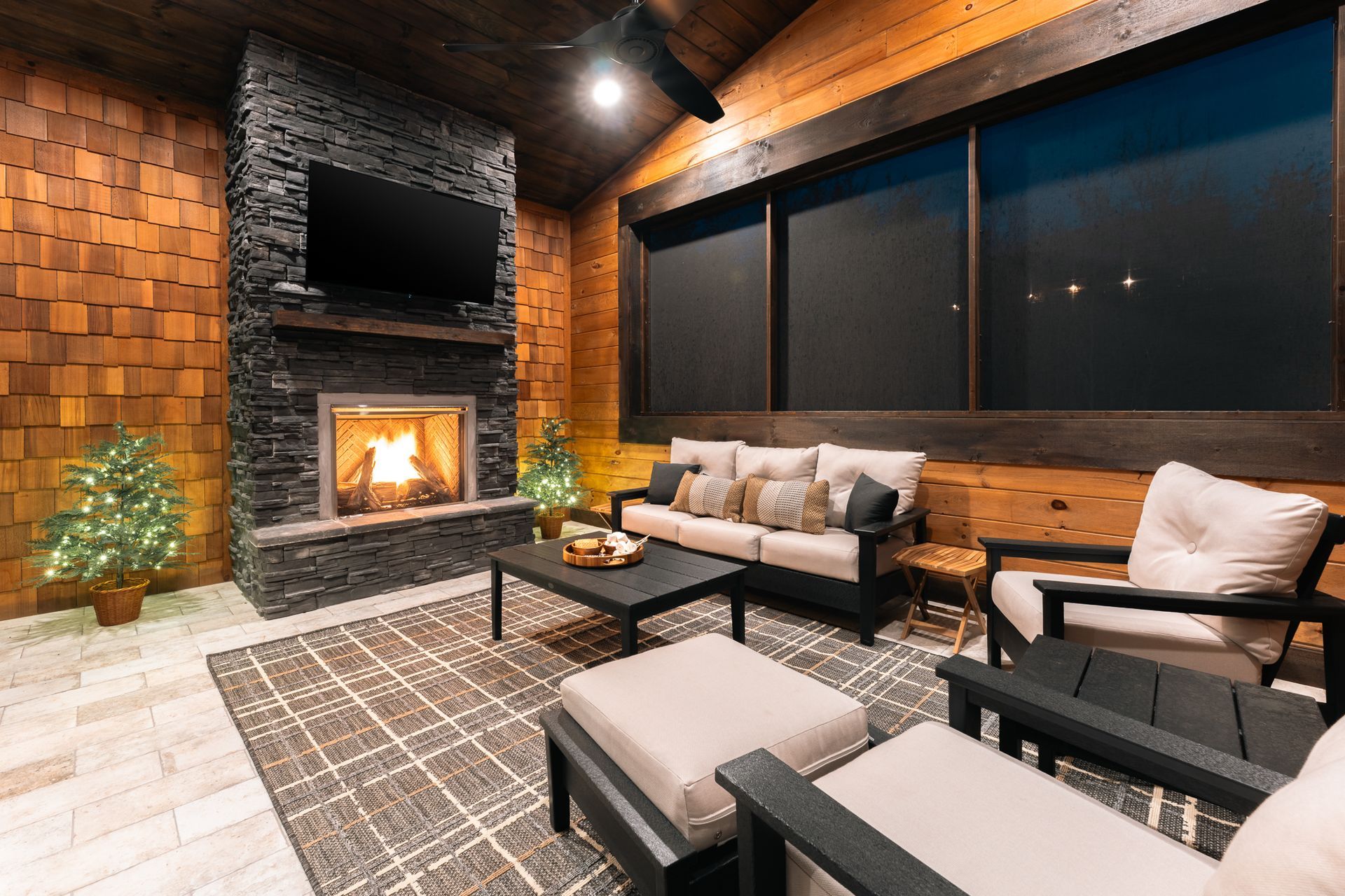 A cozy outdoor patio with a stone fireplace, television, plush seating, and a patterned rug on a wood-paneled wall room.