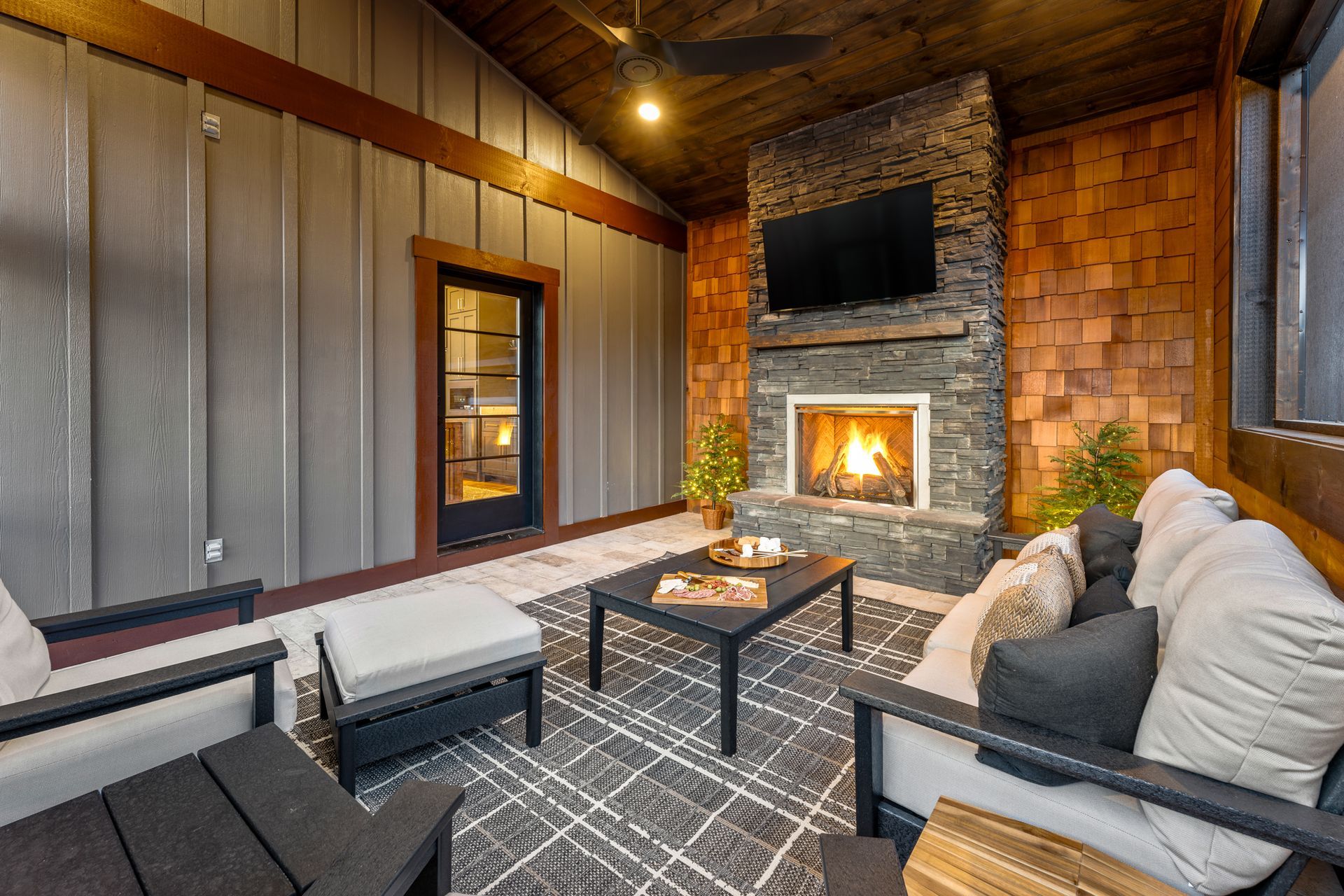 Cozy, rustic sunroom featuring a stone fireplace, dark wood ceiling, modern patio furniture, and a geometric area rug.