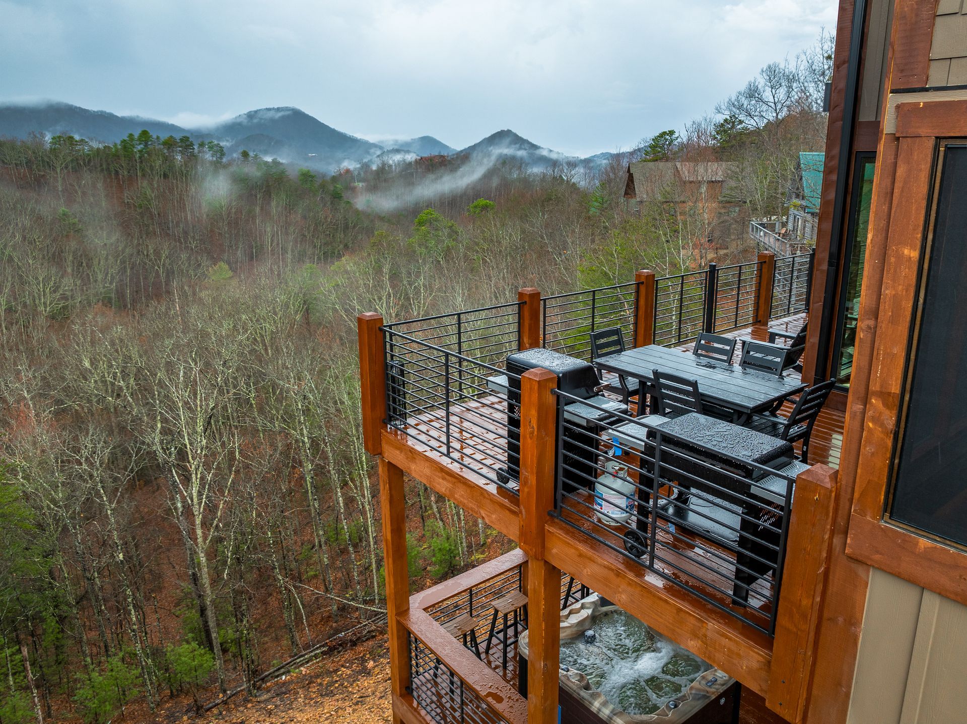 A two-story wooden deck with outdoor seating and a hot tub overlooking a misty, forested mountain landscape.