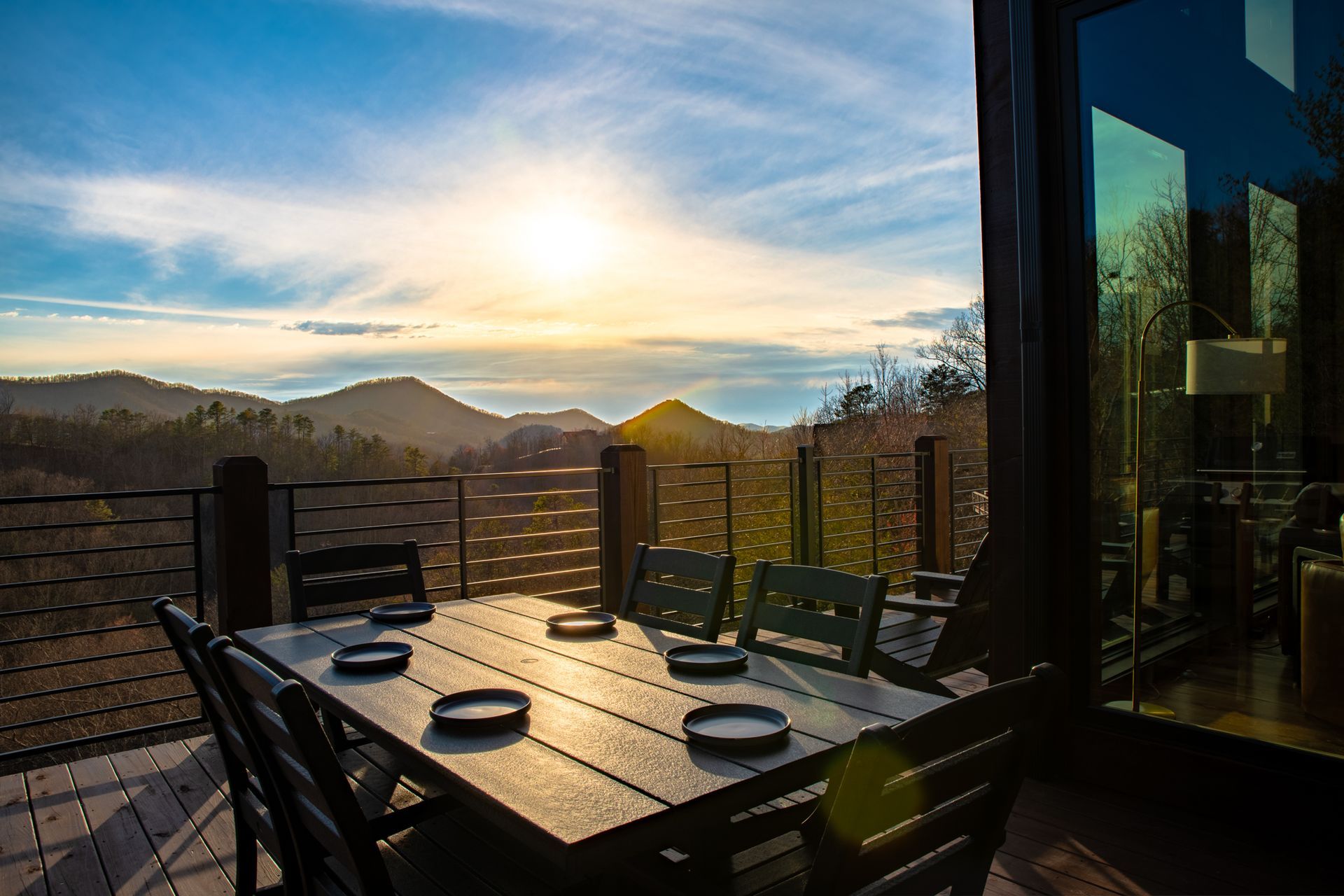 A dining table set for four on a wooden deck overlooking mountainous terrain during a bright sunset.