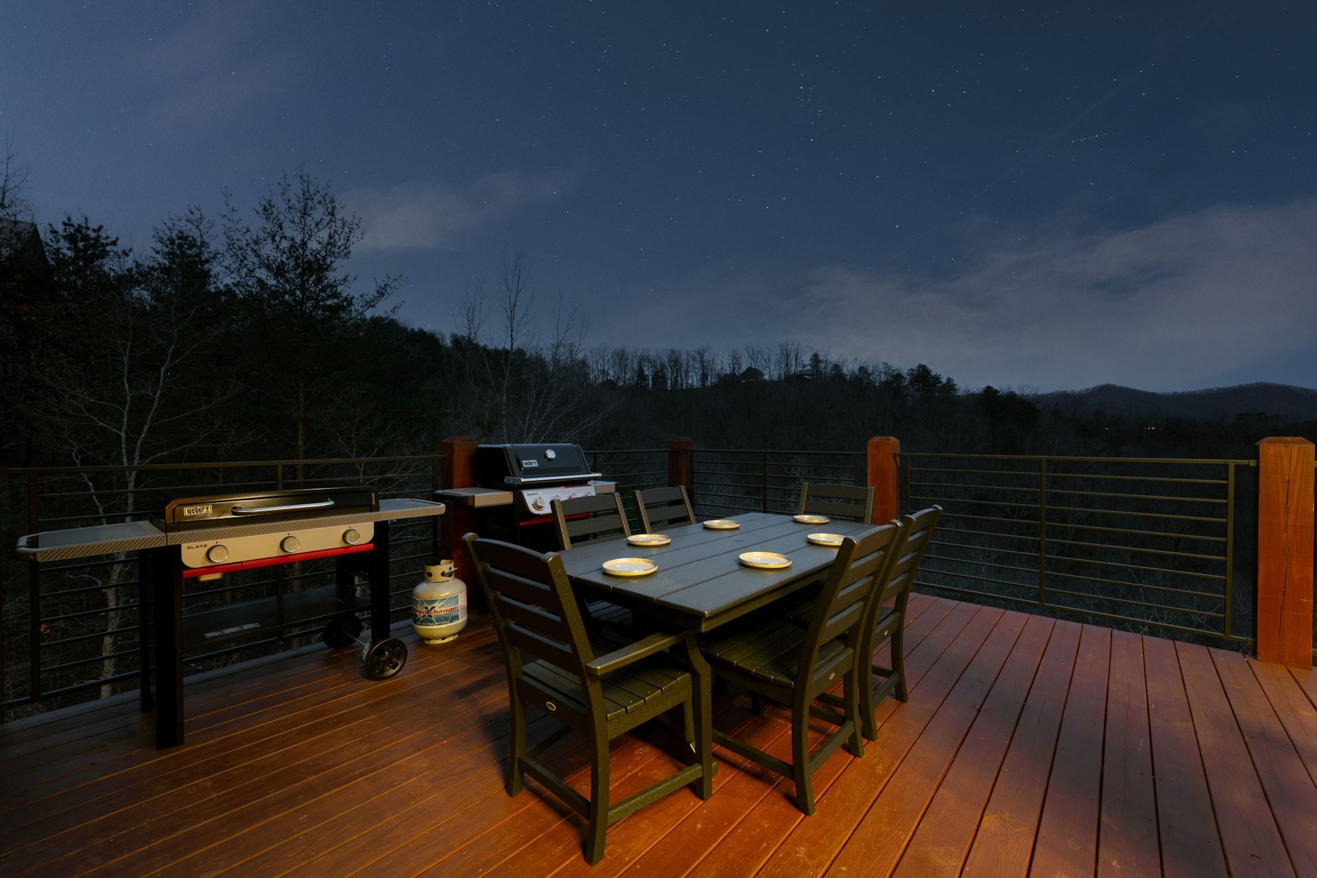 A dark wooden deck at night featuring a dining table with chairs, two grills, and a scenic mountain view under starry sky.