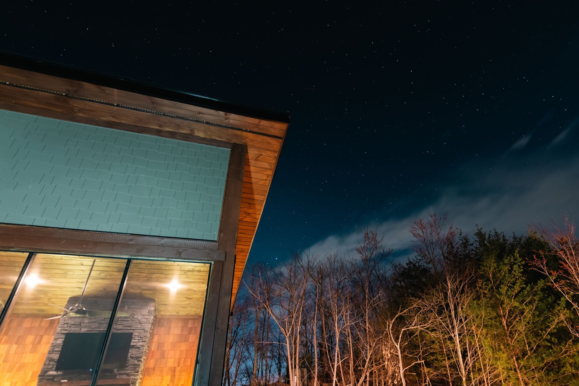 A low-angle view of a modern building with wooden eaves and a lit interior, set against a dark starry sky and tree line.